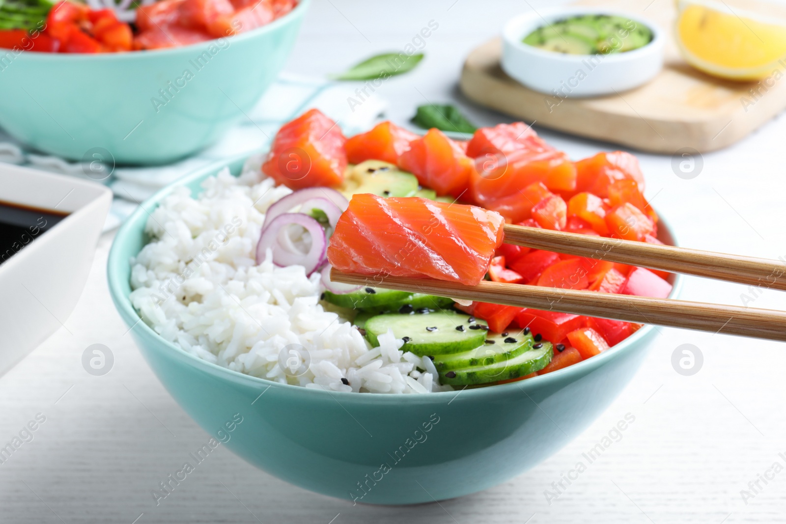 Photo of Chopsticks with piece of salmon over delicious poke bowl on white wooden table, closeup