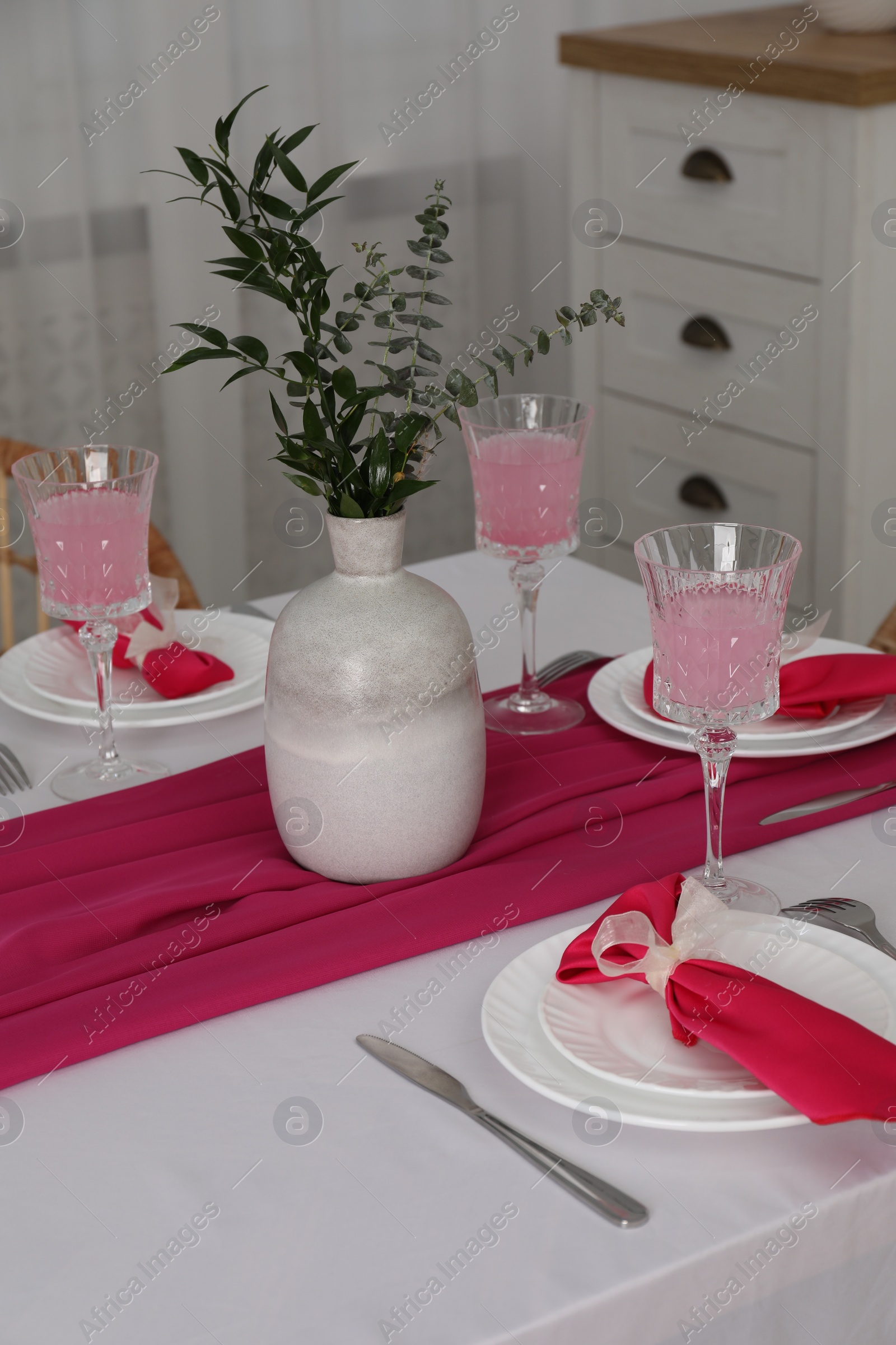 Photo of Table setting. Glasses of tasty beverage, plates, pink napkins and vase with green branches in dining room