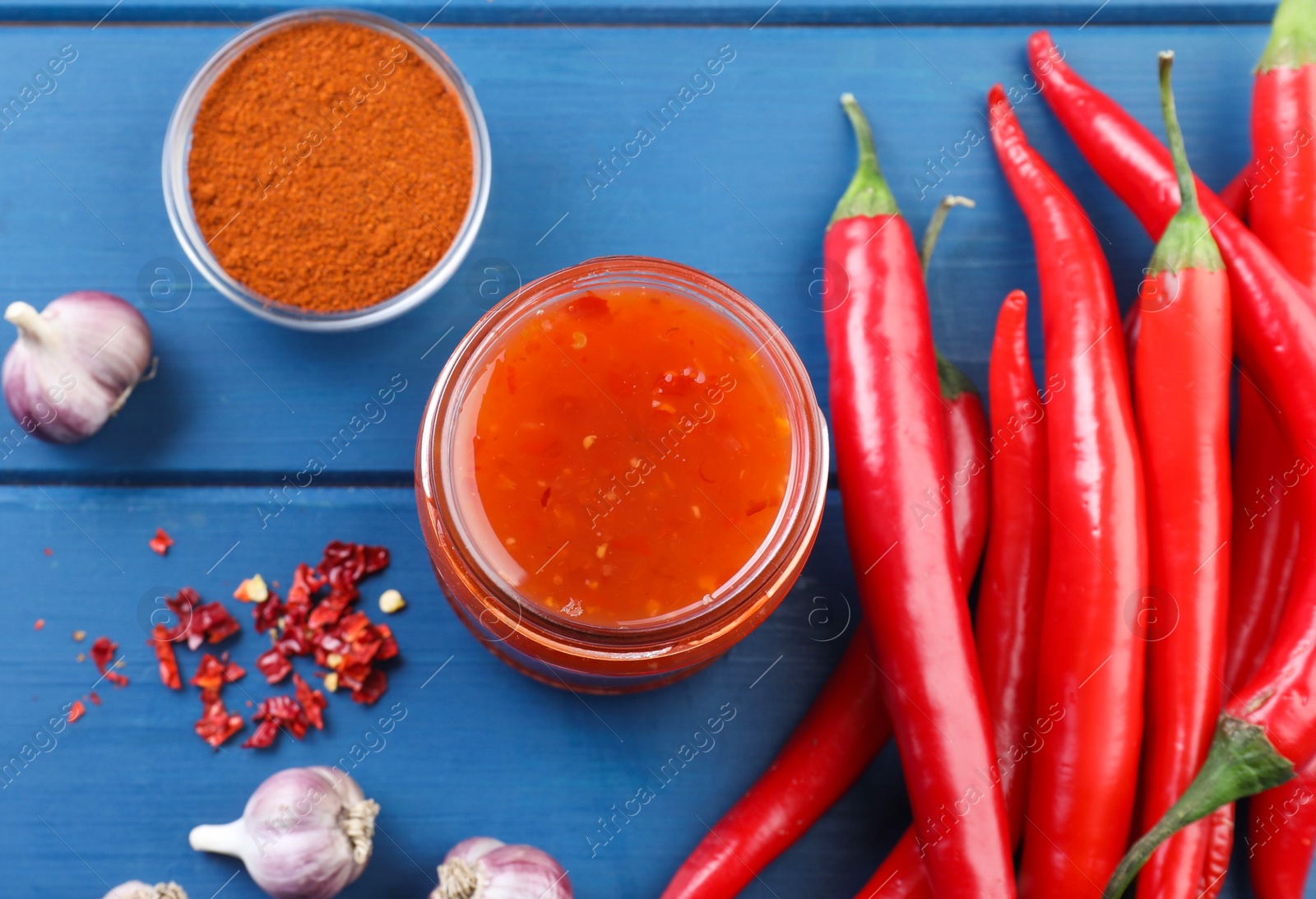 Photo of Spicy chili sauce in jar and ingredients on blue wooden table, flat lay