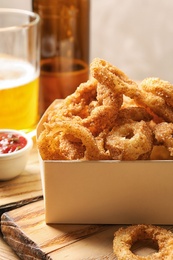 Photo of Cardboard box with crunchy fried onion rings on wooden board, closeup