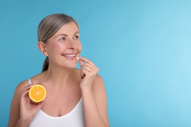 Photo of Beautiful woman with half of orange taking vitamin pill on light blue background, space for text