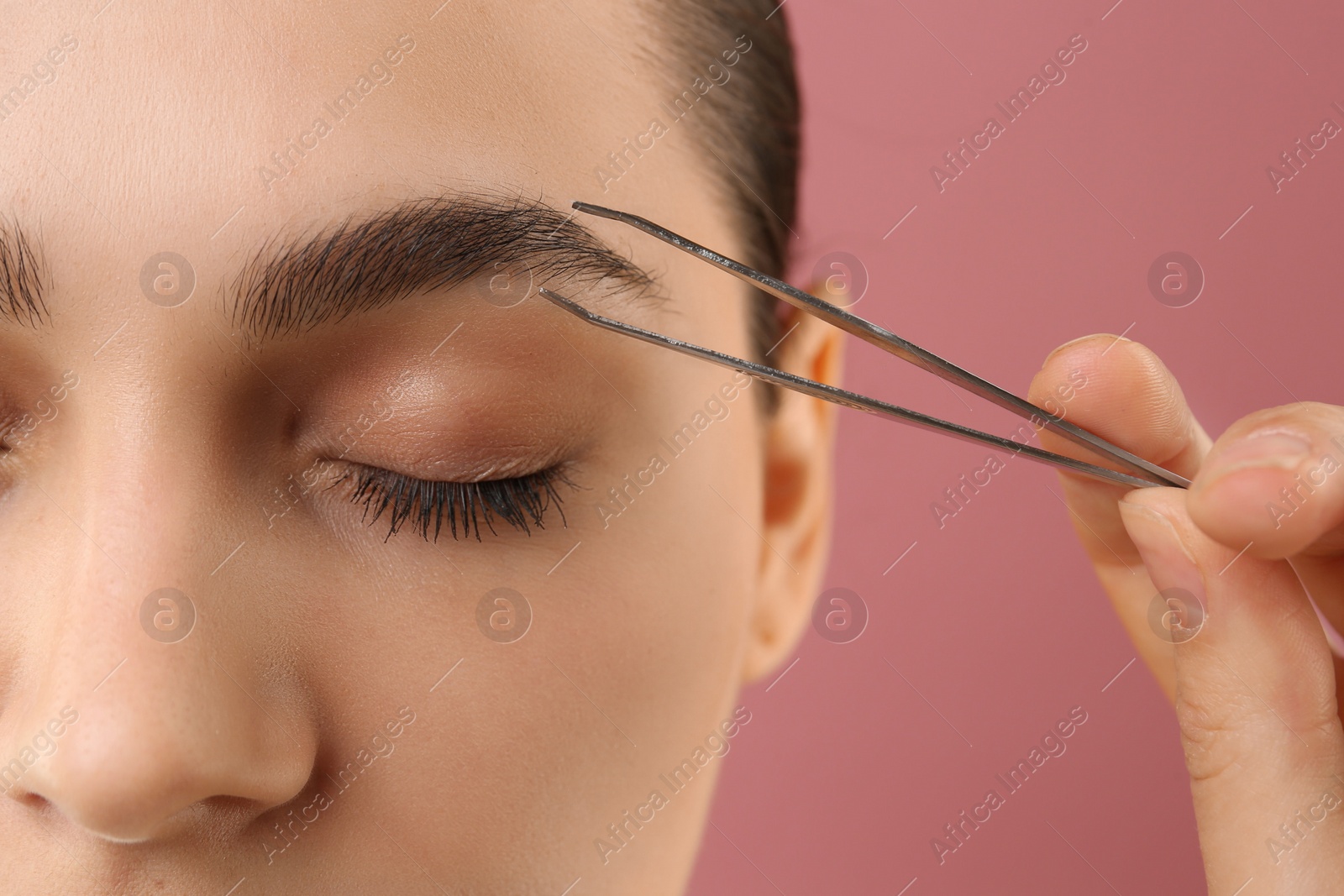 Photo of Eyebrow correction. Young woman with tweezers on pink background, closeup