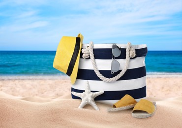 Image of Stylish bag with different accessories on sandy beach near ocean