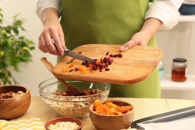 Photo of Making granola. Woman adding dried apricots and cherries into bowl with oat flakes at table in kitchen, closeup