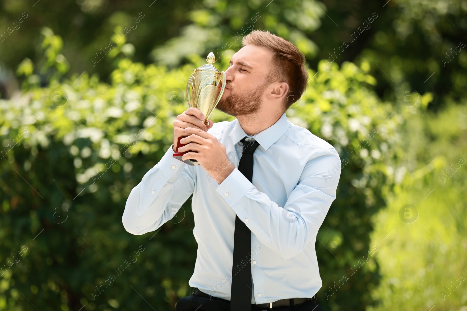 Photo of Portrait of young businessman kissing gold trophy cup in green park