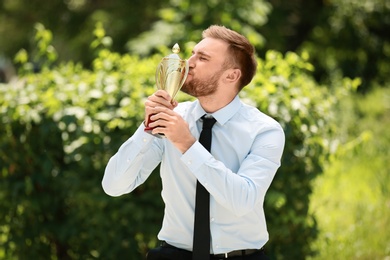 Portrait of young businessman kissing gold trophy cup in green park