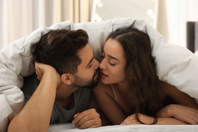 Photo of Passionate young couple kissing under warm blanket in bed indoors