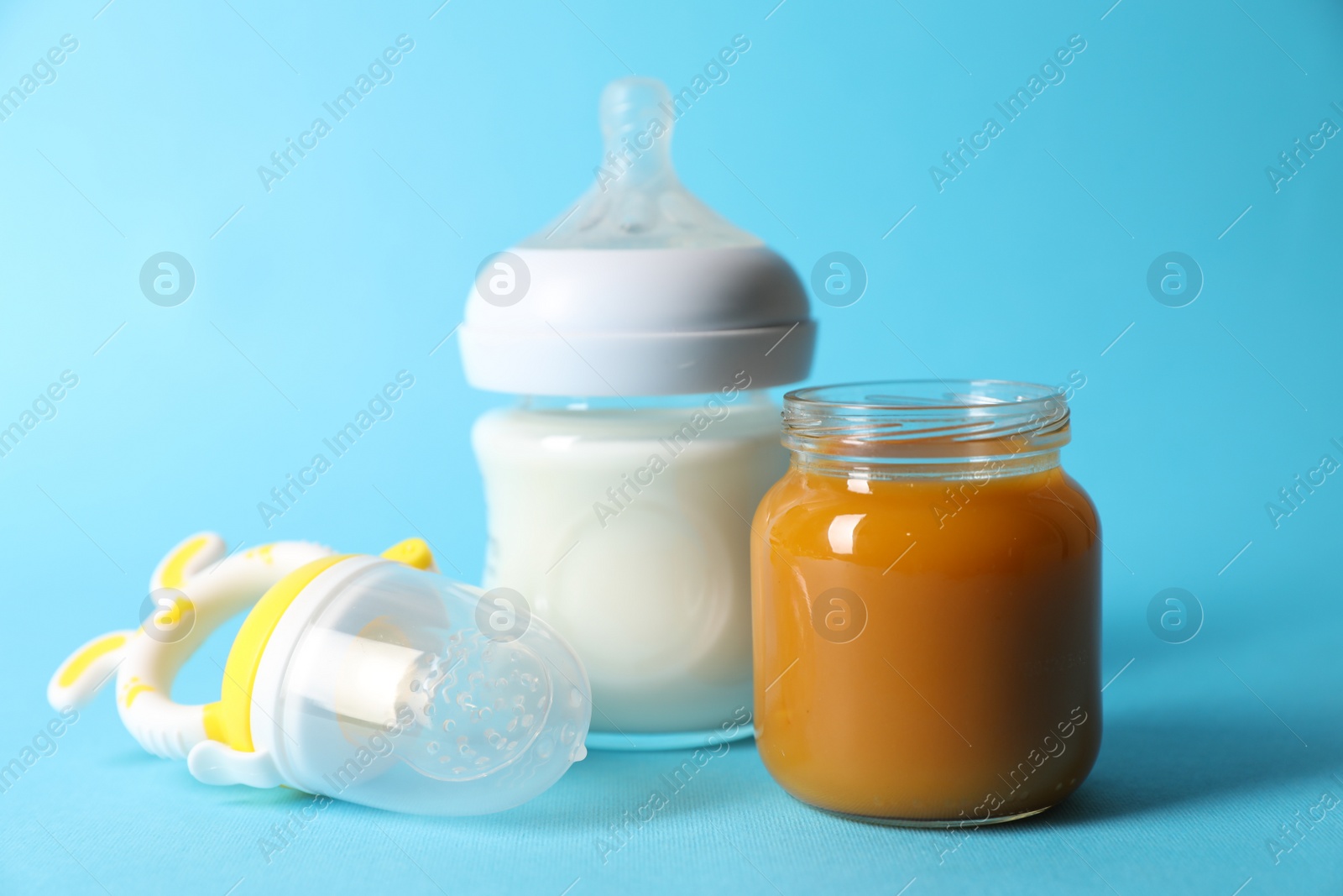 Photo of Jar with healthy baby food, bottle of milk and fruit feeder on light blue background