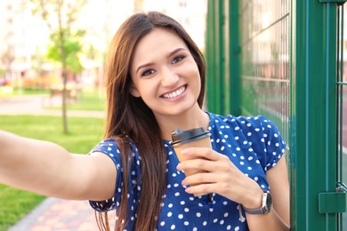 Photo of Young woman with cup of coffee taking selfie outdoors