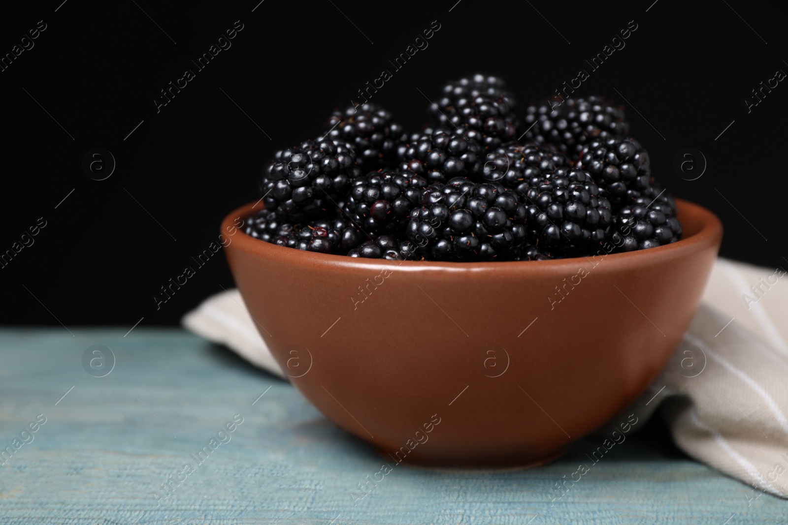 Photo of Fresh ripe blackberries in bowl on blue wooden table against dark background, closeup