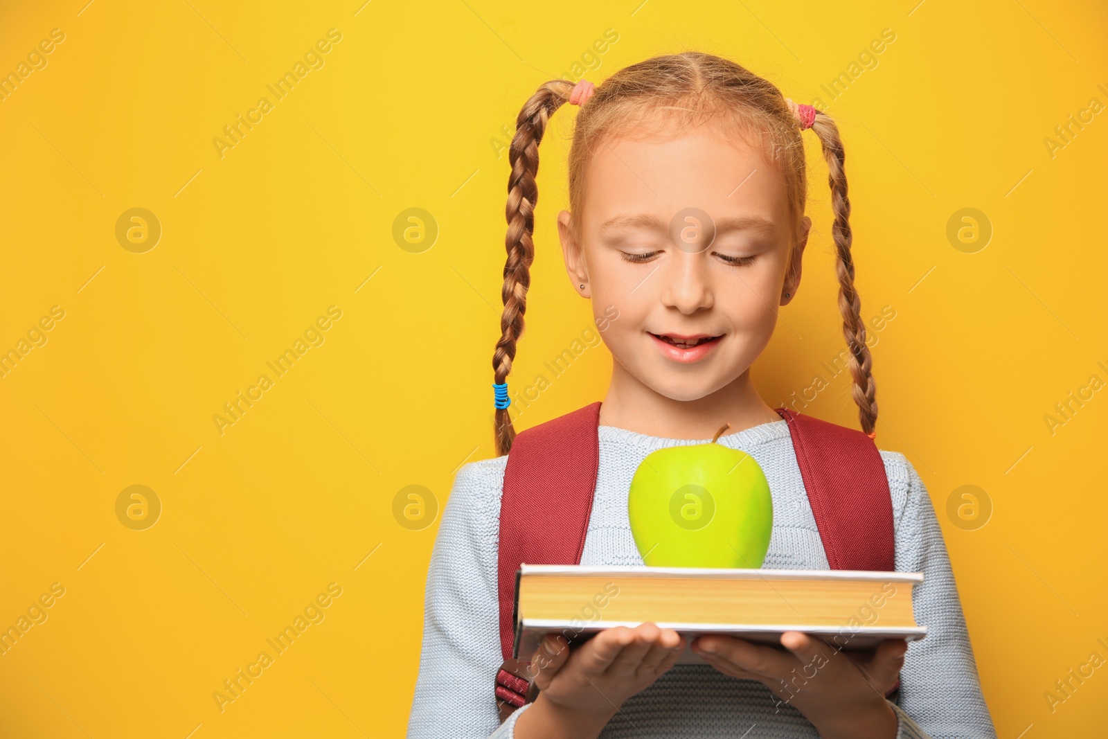 Photo of Cute little girl with apple, backpack and book on yellow background. Space for text