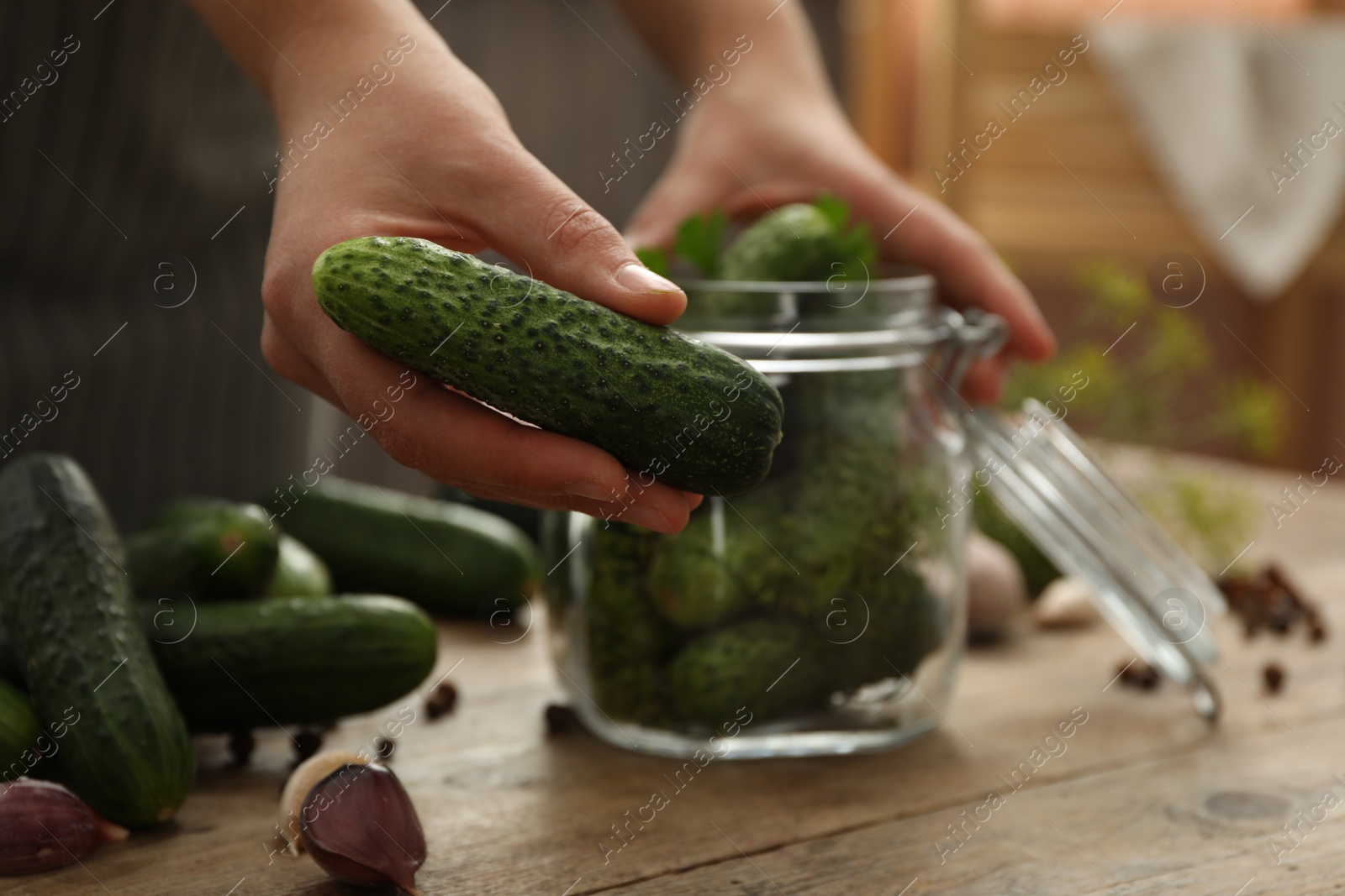 Photo of Woman putting cucumber into glass jar at wooden kitchen table, closeup. Pickling vegetables