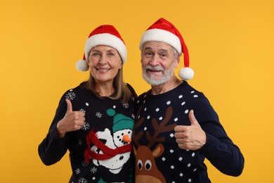 Photo of Happy senior couple in Christmas sweaters and Santa hats showing thumbs up on orange background