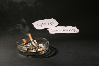 Ashtray with burnt cigarettes and words Stop Smoking written on paper on black table