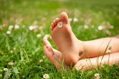 Photo of Child with chamomile between toes sitting on green grass outdoors, closeup of feet