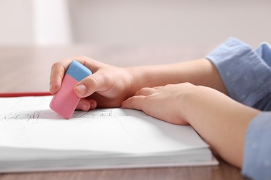 Girl erasing drawing in her book at wooden table indoors, closeup