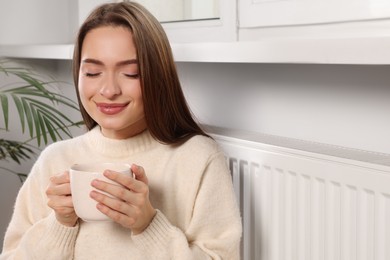 Photo of Woman holding cup with hot drink near heating radiator indoors