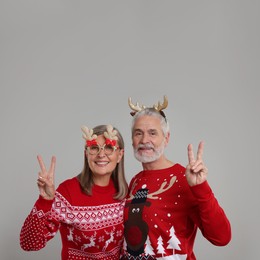 Photo of Senior couple in Christmas sweaters, reindeer headband and funny glasses showing V-sign on grey background