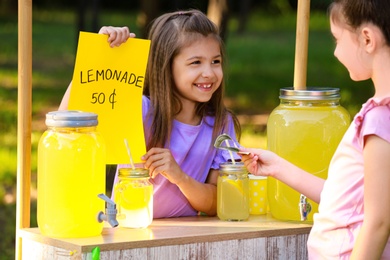 Photo of Cute little girl selling natural lemonade to kid in park. Summer refreshing drink