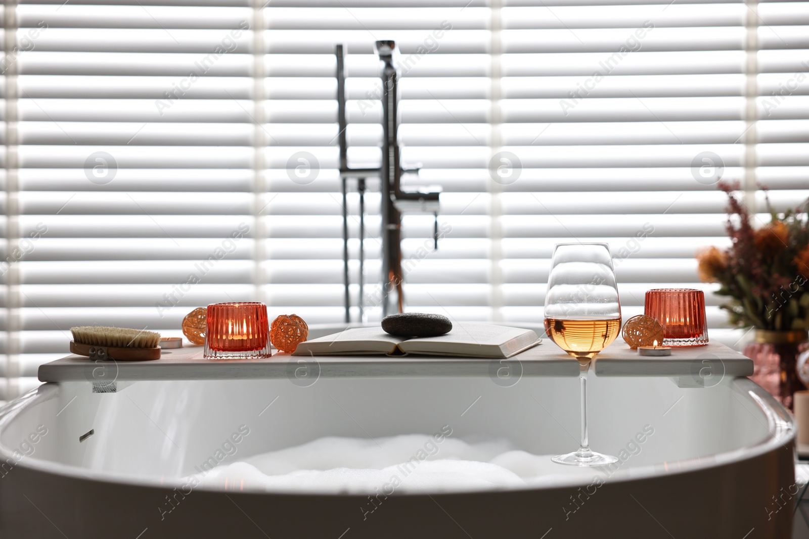 Photo of White wooden tray with glass of rose wine, book and burning candles on bathtub in bathroom