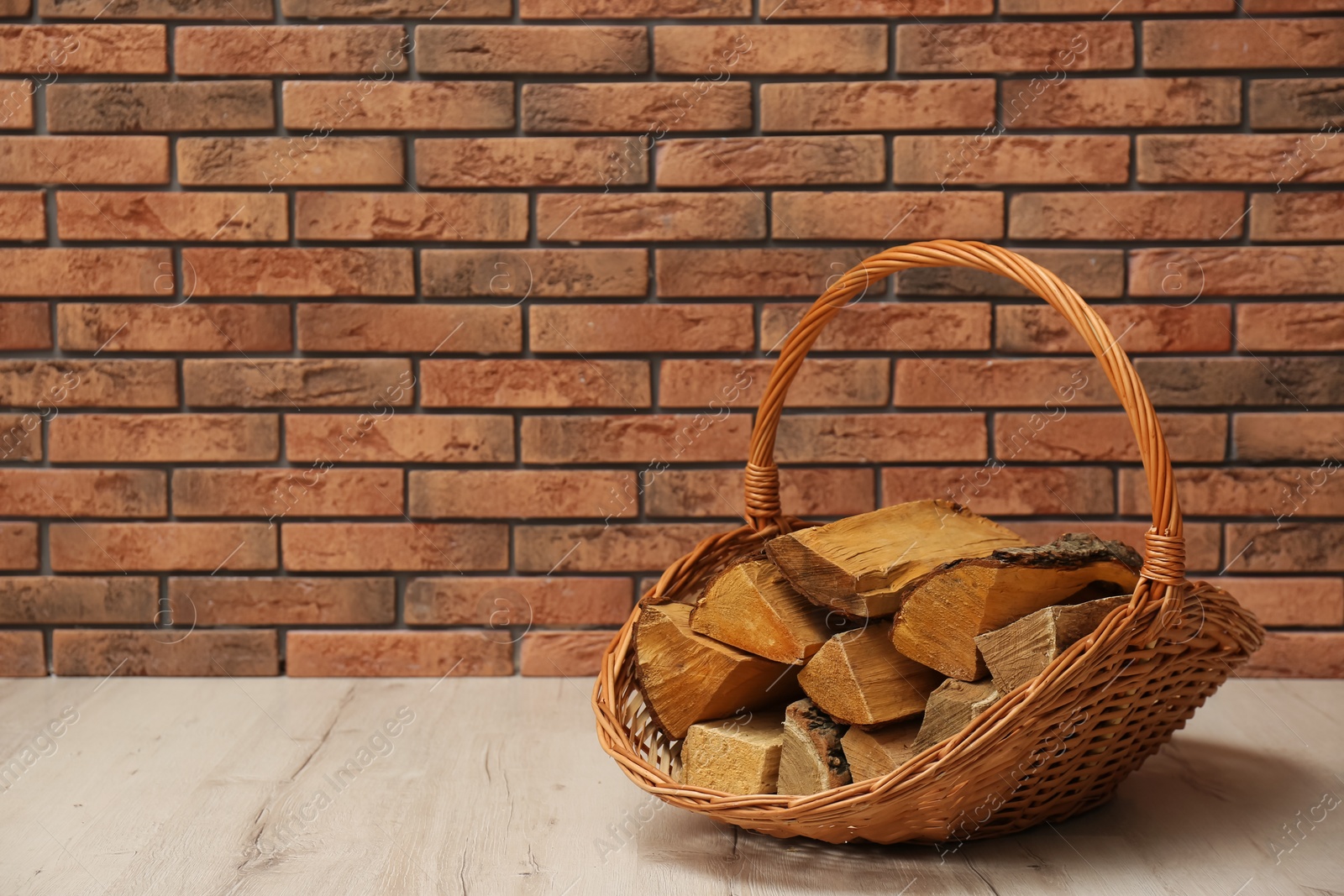 Photo of Wicker basket with firewood near brick wall indoors, space for text