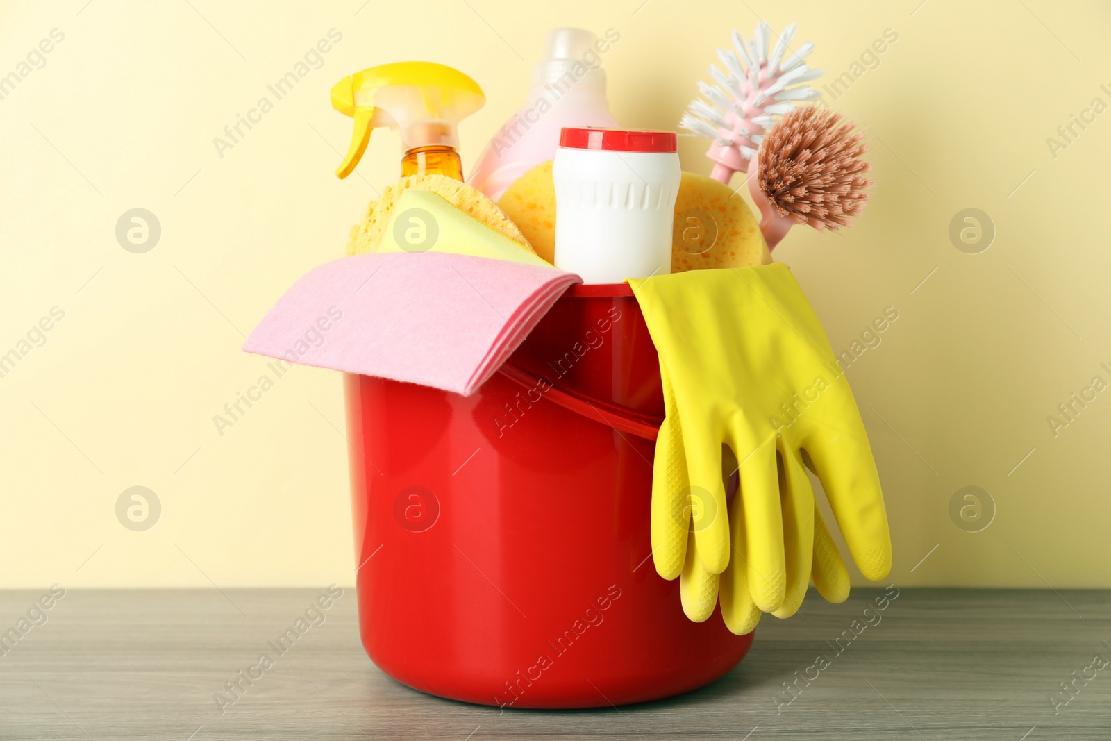 Photo of Bucket with different cleaning supplies on wooden floor near beige wall, closeup