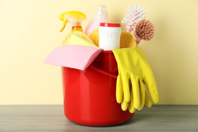 Photo of Bucket with different cleaning supplies on wooden floor near beige wall, closeup