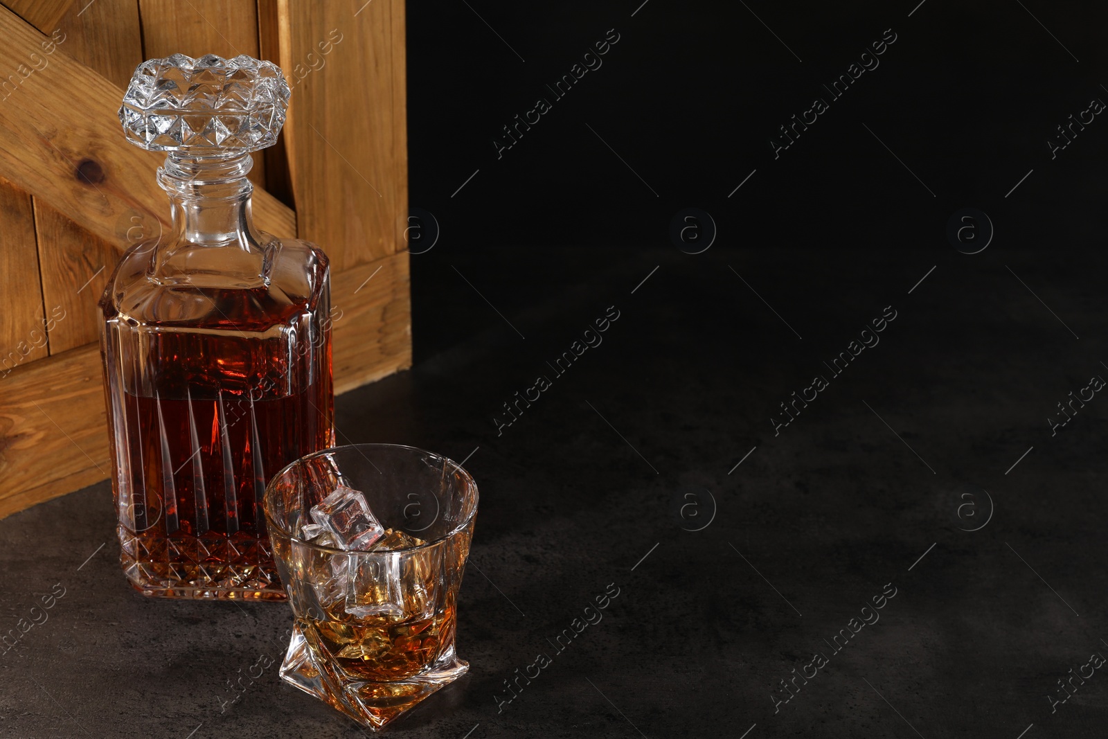 Photo of Whiskey in glass and bottle near wooden crate on dark table against black background. Space for text