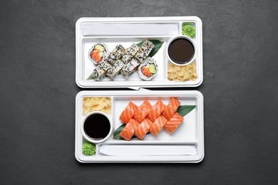 Photo of Plastic containers with delicious sushi rolls, soy sauce, ginger, wasabi and chopsticks on black table, flat lay. Food delivery