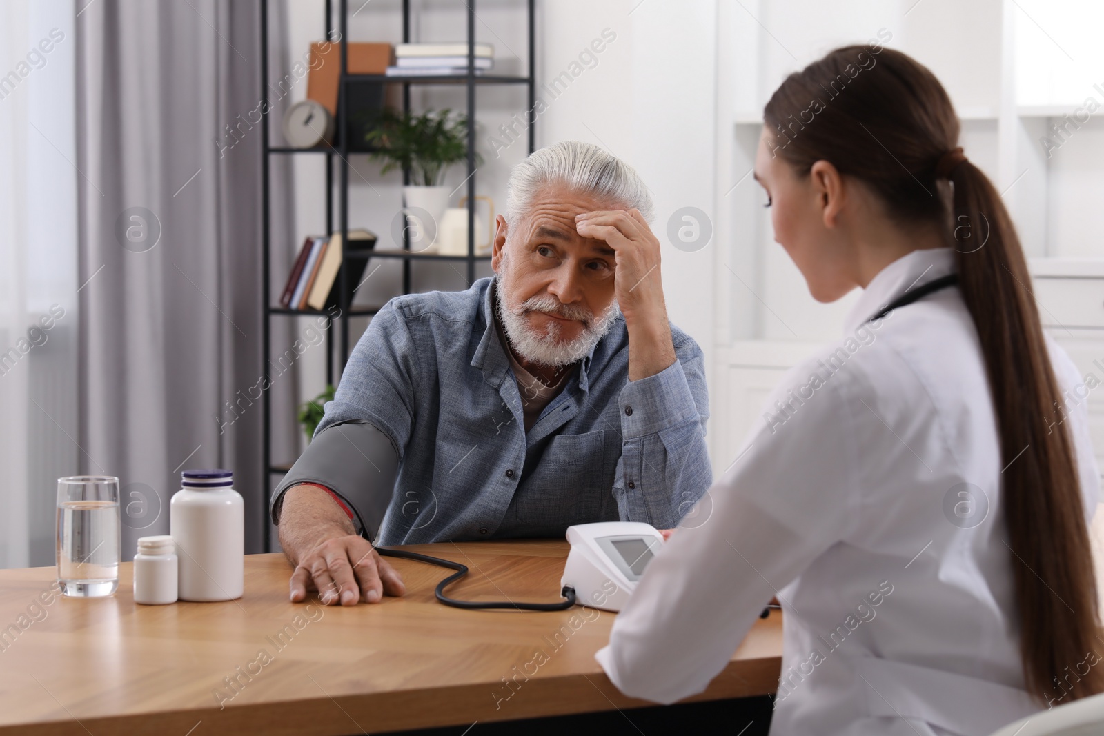 Photo of Young healthcare worker measuring senior man's blood pressure at wooden table indoors