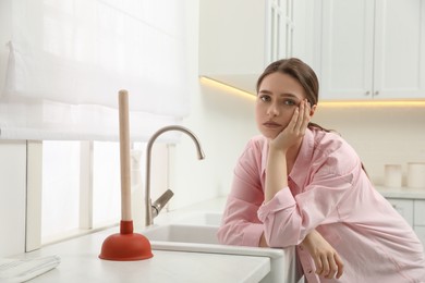 Photo of Unhappy young woman with plunger near clogged sink in kitchen
