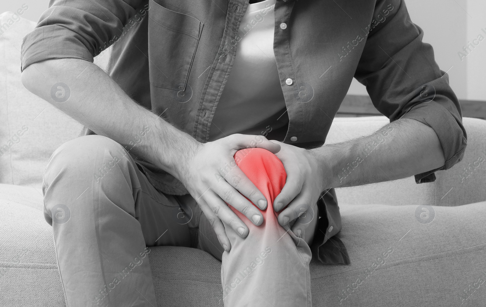 Image of Man suffering from pain in knee on sofa at home, closeup. Black and white effect