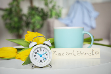 Photo of Alarm clock, aromatic morning coffee, beautiful flowers and card with RISE AND SHINE wish on white table indoors