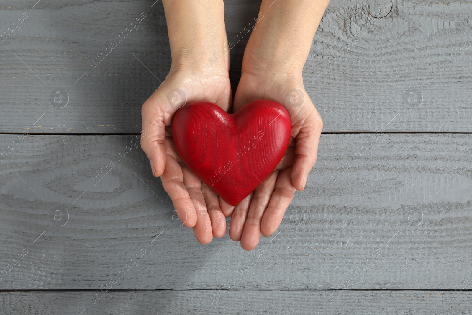 Photo of Elderly woman holding red heart in hands at grey wooden table, top view