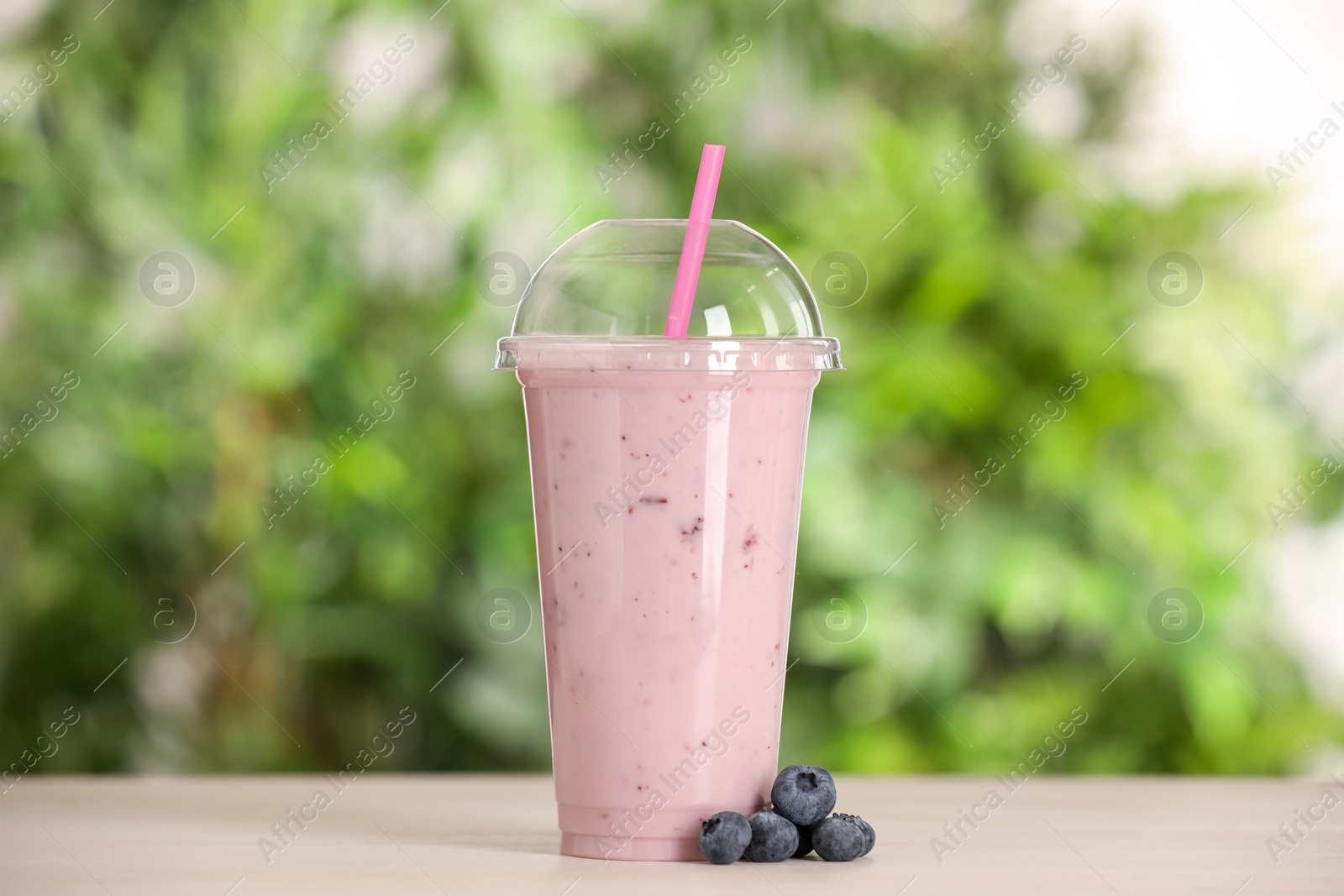 Photo of Plastic cup of tasty smoothie and fresh blueberries on wooden table outdoors