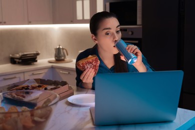 Photo of Young woman eating sandwich and drinking soda while using laptop in kitchen at night. Bad habit