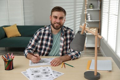 Young man coloring antistress picture at table indoors