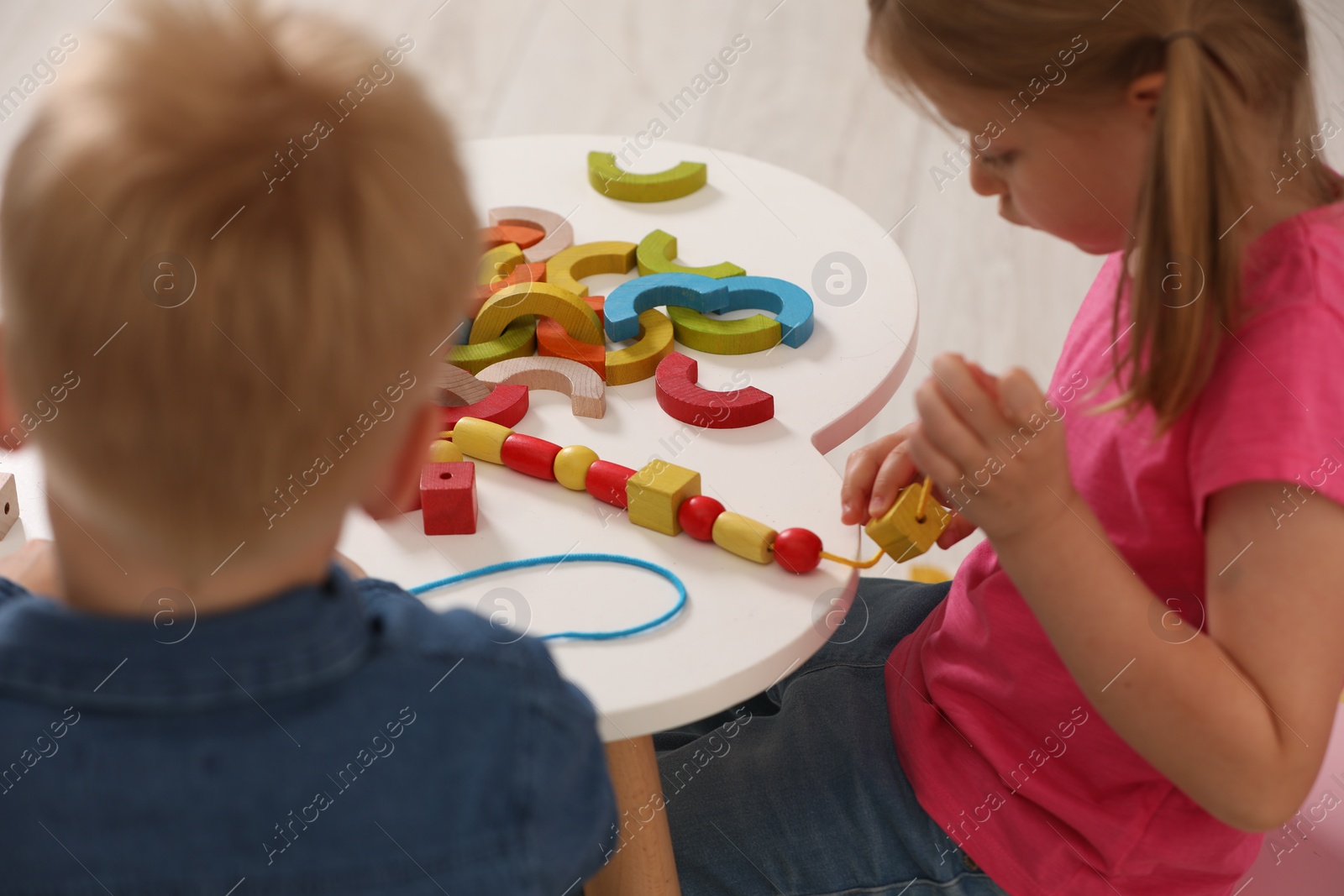 Photo of Little children playing with wooden pieces and string for threading activity at white table indoors. Developmental toys