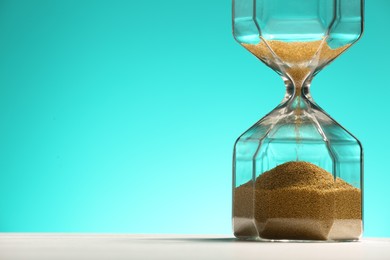 Photo of Hourglass with flowing sand on white table against turquoise background, closeup. Space for text