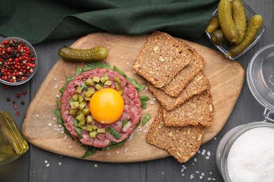 Photo of Tasty beef steak tartare served with yolk, pickled cucumber and other accompaniments on grey wooden table, flat lay