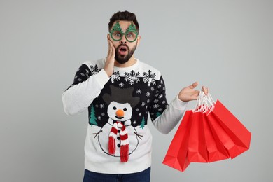 Shocked young man in Christmas sweater and funny glasses with shopping bags on grey background