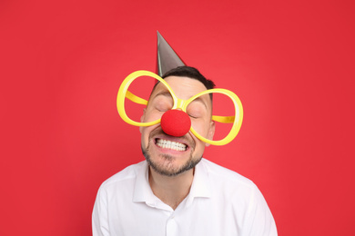 Photo of Funny man with large glasses, party hat and clown nose on red background. April fool's day