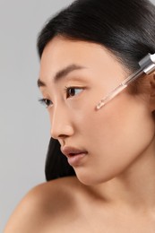 Photo of Beautiful young woman applying cosmetic serum onto her face on grey background, closeup