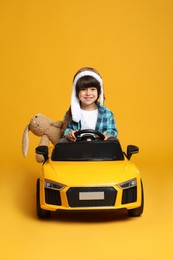 Photo of Cute little boy with toy bunny driving children's car on yellow background