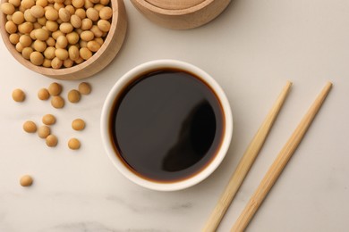 Photo of Soy sauce in bowl, soybeans and chopsticks on white marble table, flat lay