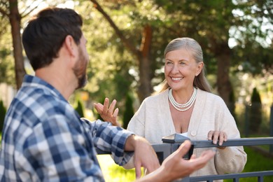 Photo of Friendly relationship with neighbours. Happy senior woman talking to man near fence outdoors