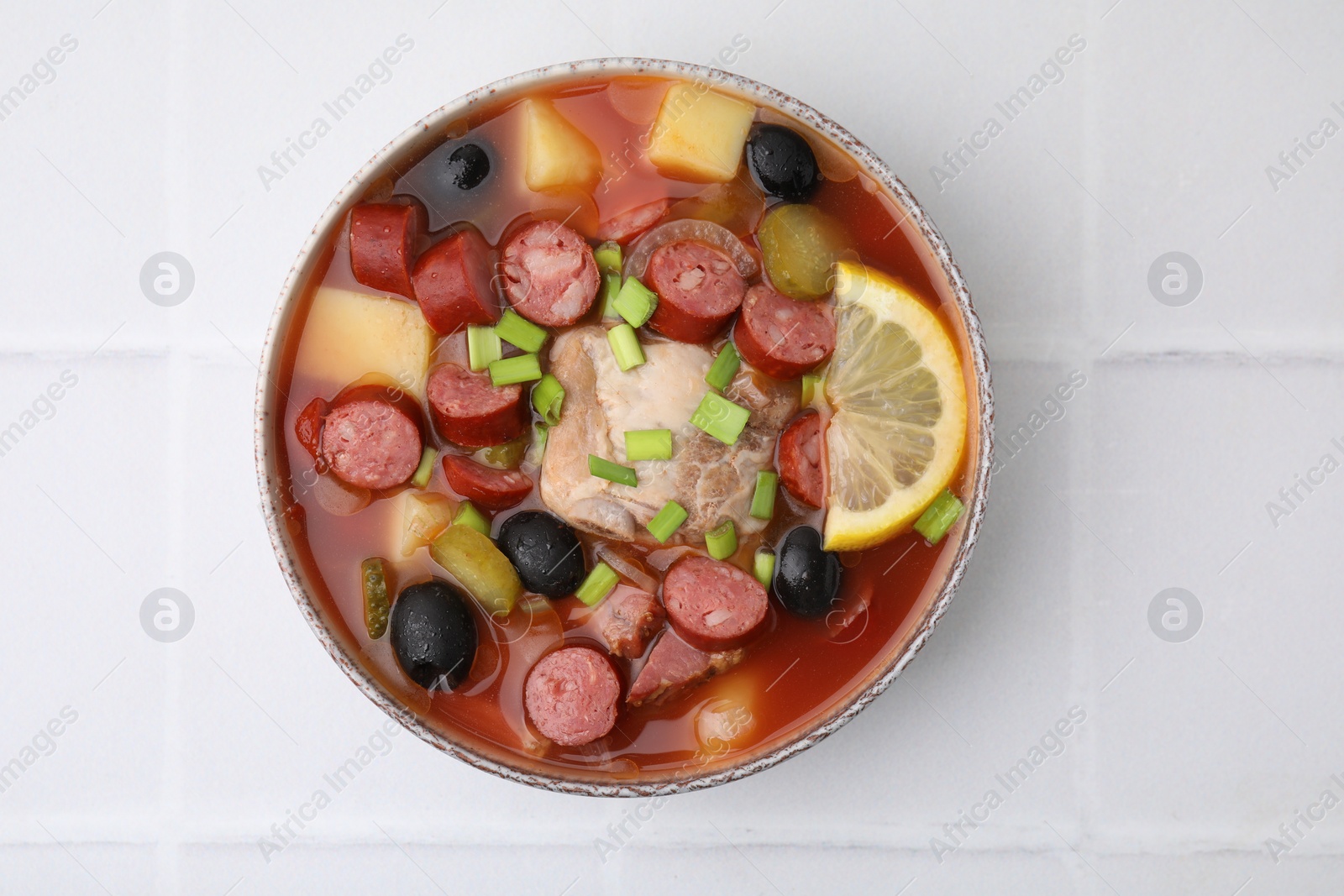 Photo of Meat solyanka soup with thin dry smoked sausages in bowl on white tiled table, top view