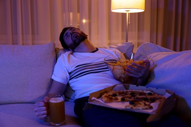 Photo of Man with chips, pizza and glassbeer sleeping on sofa at night. Bad habit