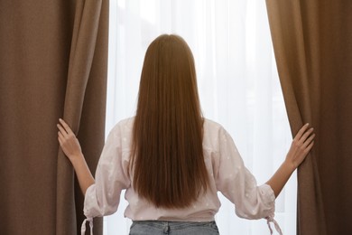 Woman opening elegant window curtains in room, back view