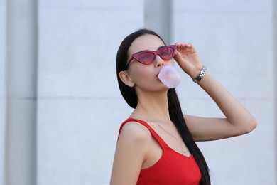 Beautiful woman in stylish sunglasses blowing gum near wall outdoors